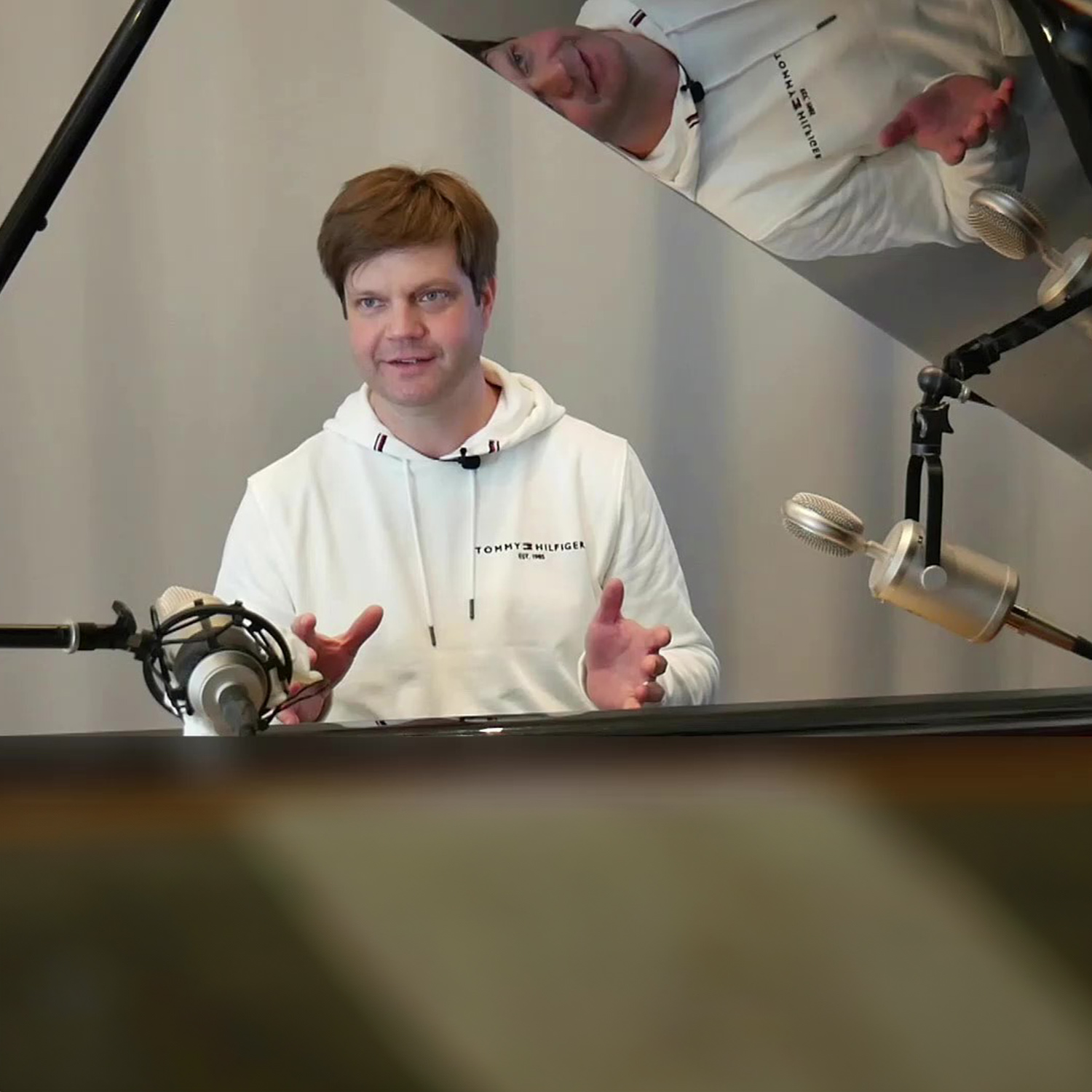 Person im weißen Hoodie spricht, umgeben von Mikrofonen und reflektiertem Klavierdeckel.