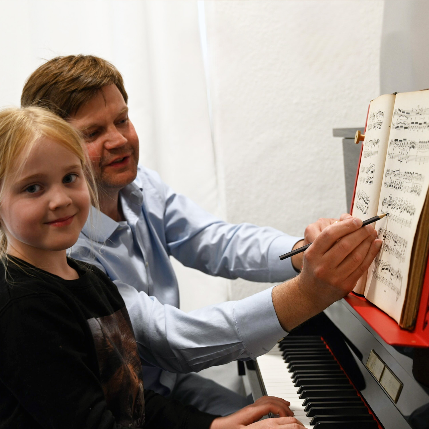 Mann zeigt Kind am Klavier mit Bleistift auf Notenblatt; beide lächeln.