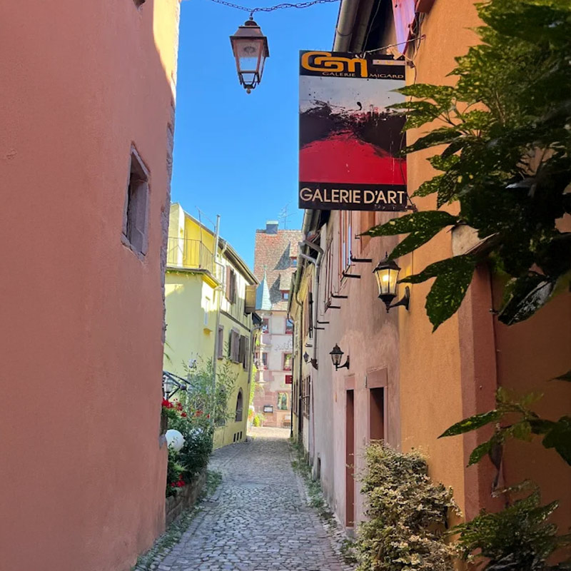 Schmale, gepflasterte Gasse mit bunten Häusern, Galerieschild und Pflanzen; klarer Himmel.
