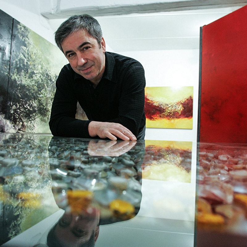 Mann in dunkler Kleidung lehnt sich über glastisch, Gemälde an der Wand, bunte Steine unter Glas.