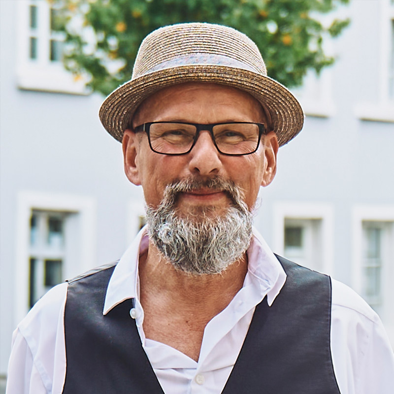 Mann mit Brille und Strohhut, weißem Hemd und Weste, lächelt, Hintergrund unscharf mit Gebäude und Baum.